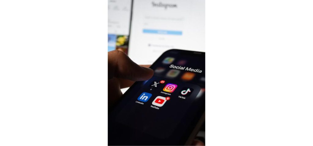 Person holding a smartphone showing TikTok, Instagram, X, LinkedIn and YouTube app icons under a folder labeled Social Media